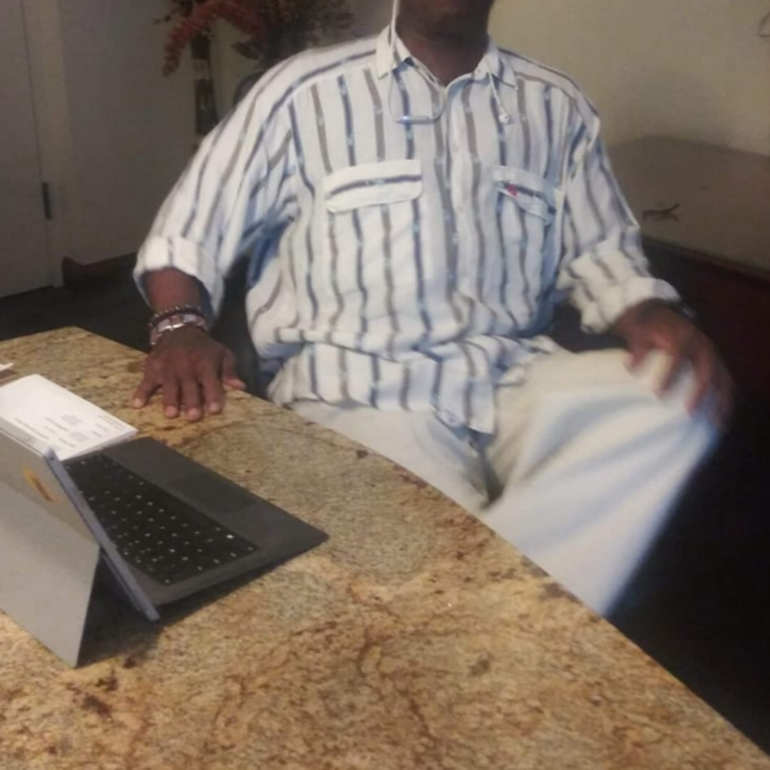 Man seated at a granite table with a laptop in front of him, wearing a striped shirt and light pants.