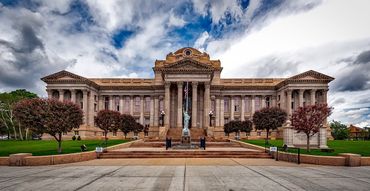 Grand neoclassical building with columns and red-topped trees under a cloudy sky