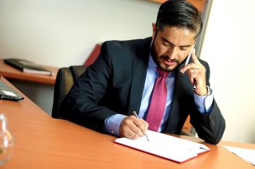 Businessperson in a dark suit talking on the phone while writing on paper at a desk.